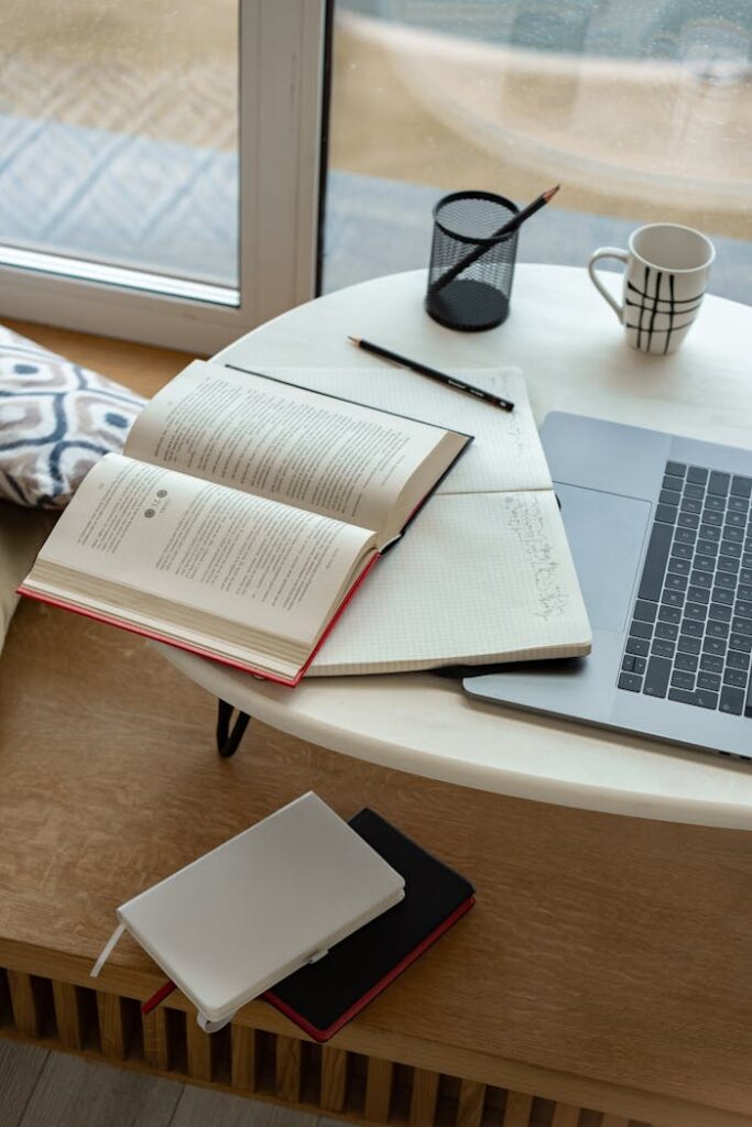 pexels-photo-7583936 A cozy indoor study setup featuring an open book, laptop, and coffee mug by the window.